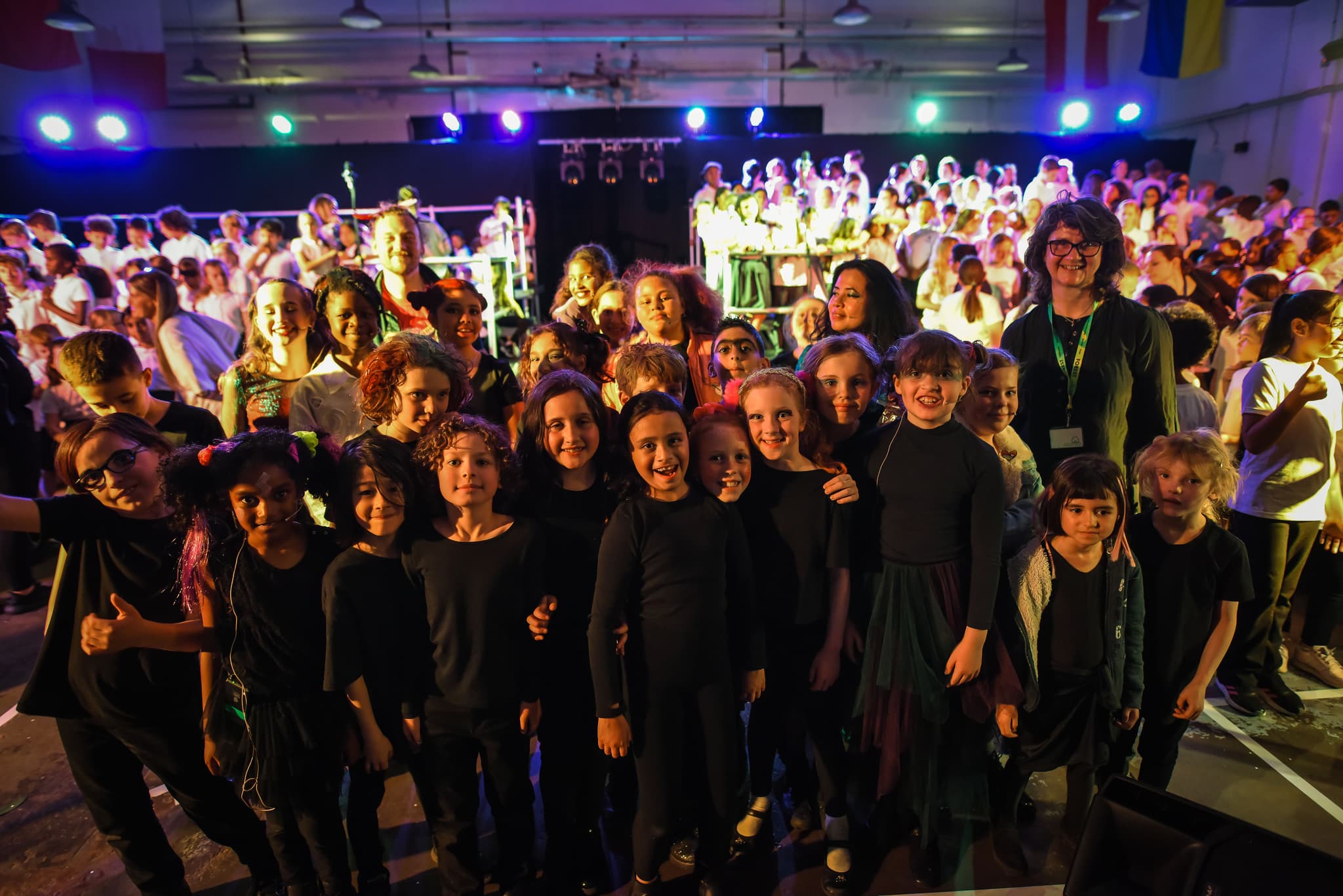 A group of children and an adult stand smiling in front of a large choir on a brightly lit stage.