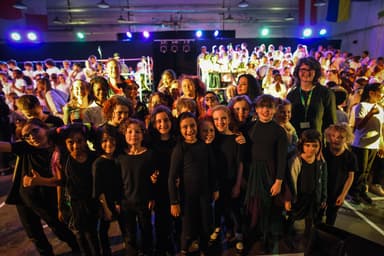 A group of children and an adult stand smiling in front of a large choir on a brightly lit stage.
